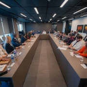 Foto geral de uma sala com uma grande mesa em formato de U, de cor marrom. Há mais de 20 pessoas sentadas junto à mesa. O ambiente é formal.