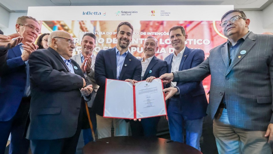 Um grupo de sete homens, vestidos de terno ou blazer, posam para uma foto em um evento. Um deles, com barba, no centro da imagem, sorri enquanto segura um documento de capa vermelha e branca. Outro homem, mais velho e de óculos, à sua esquerda, parece aplaudir. Os demais estão em volta, observando e sorrindo. O cenário, desfocado, parece ser o de um palco com a iluminação direcionada para as pessoas. Ao fundo, pode-se ler os nomes InBetta, Sapucaia do Sul, e Rio Grande do Sul em painéis.
