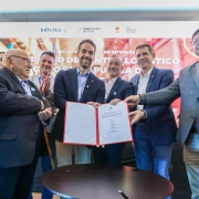Um grupo de sete homens, vestidos de terno ou blazer, posam para uma foto em um evento. Um deles, com barba, no centro da imagem, sorri enquanto segura um documento de capa vermelha e branca. Outro homem, mais velho e de óculos, à sua esquerda, parece aplaudir. Os demais estão em volta, observando e sorrindo. O cenário, desfocado, parece ser o de um palco com a iluminação direcionada para as pessoas. Ao fundo, pode-se ler os nomes InBetta, Sapucaia do Sul, e Rio Grande do Sul em painéis.