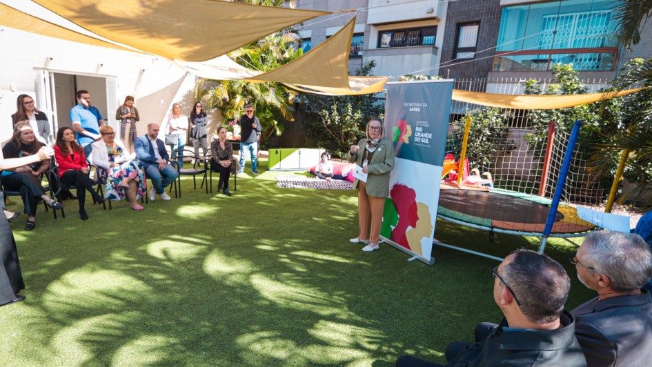 Em um pátio externo com gramado sintético, cerca de 20 pessoas participam de um evento. À direita, uma mulher de cabelos claros e óculos fala em pé, segurando papéis, ao lado de um banner com a identidade visual da Secretaria da Saúde do Governo do Rio Grande do Sul. À esquerda, o público acompanha sentado e em pé; acima, toldos triangulares bege sombreiam o espaço, criando desenhos de sombra no chão. Ao fundo há plantas, brinquedos infantis e uma cama elástica cercada por rede colorida, com fachadas de prédios residenciais compondo o cenário.