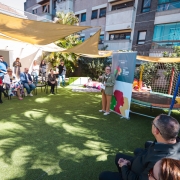 Em um pátio externo com gramado sintético, cerca de 20 pessoas participam de um evento. À direita, uma mulher de cabelos claros e óculos fala em pé, segurando papéis, ao lado de um banner com a identidade visual da Secretaria da Saúde do Governo do Rio Grande do Sul. À esquerda, o público acompanha sentado e em pé; acima, toldos triangulares bege sombreiam o espaço, criando desenhos de sombra no chão. Ao fundo há plantas, brinquedos infantis e uma cama elástica cercada por rede colorida, com fachadas de prédios residenciais compondo o cenário.