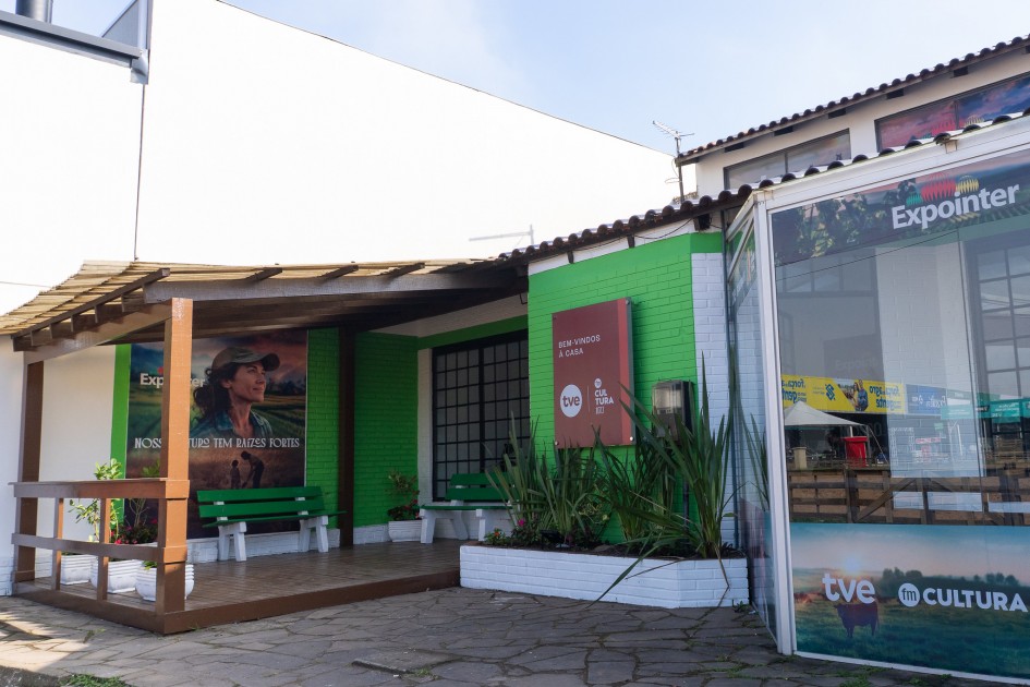 Entrada charmosa de uma casa com paredes verdes e brancas, decorada com flores e placas coloridas. Um grande letreiro vermelho dá boas-vindas à “Casa TVE Cultura”. Ao lado, um cartaz mostra uma mulher com boné diante da paisagem rural, com a frase: “Nosso futuro tem diversas cores”.