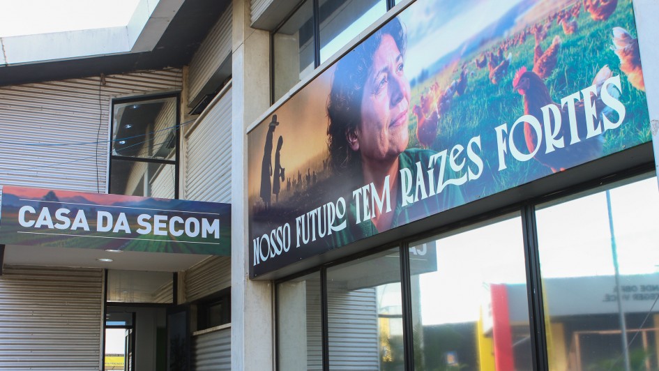 Fachada de prédio com o nome “Casa da Secom”. Um grande banner mostra cenas rurais com galinhas, pessoas no campo e o rosto de uma mulher, acompanhado da frase: “Nosso futuro tem raízes fortes”. O ambiente transmite conexão com o campo e comunicação pública.