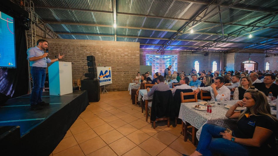 Gabriel Souza está à esquerda da foto, sobre um palco preto. Ele está de pé, discursando com um microfone na mão esquerda e gesticulando com a outra mão. Ele veste uma calça jeans e uma camisa azul claro. Ele fala para uma plateia que está sentada ao redor de mesas compridas. As pessoas olham atentas. A cena se passa em um local com teto de zinco e piso marrom.