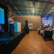 Gabriel Souza está à esquerda da foto, sobre um palco preto. Ele está de pé, discursando com um microfone na mão esquerda e gesticulando com a outra mão. Ele veste uma calça jeans e uma camisa azul claro. Ele fala para uma plateia que está sentada ao redor de mesas compridas. As pessoas olham atentas. A cena se passa em um local com teto de zinco e piso marrom. 