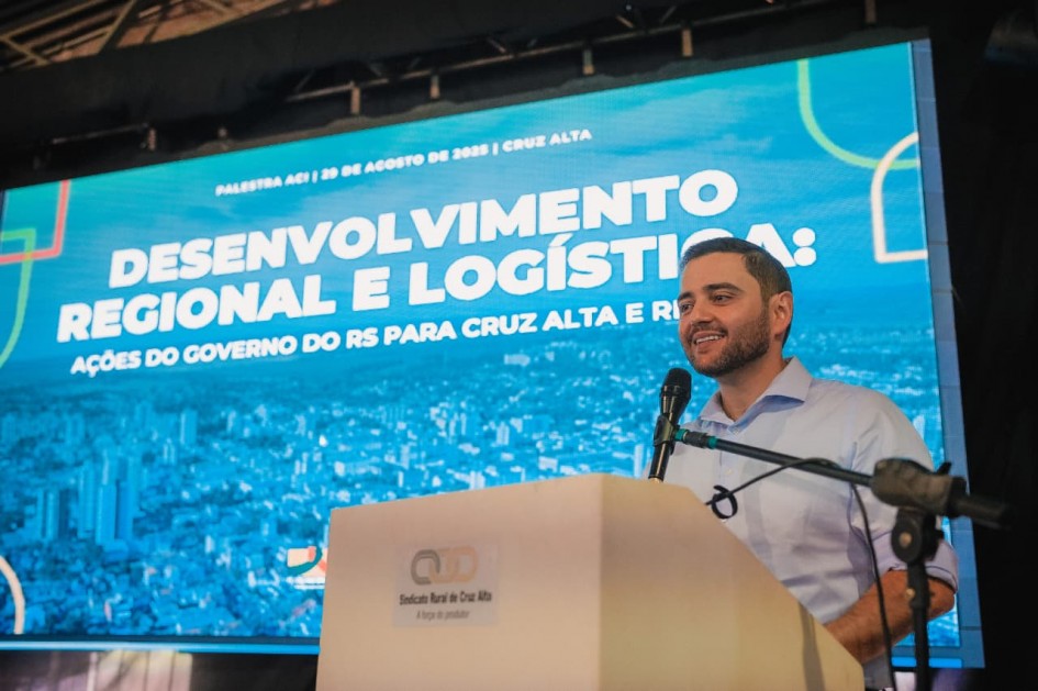 Gabriel Souza discursa atrás de um púlpito claro. Há um pedestal que segura o microfone. Gabriel está com uma camisa clara azul. Ele está em frente a um painel de vídeo onde aparece uma apresentação com imagem de fundo em azul esverdeado com uma cidade em silhueta e com um título "desenvolvimento regional e logística, ações do governo do RS para Cruz Alta e região".