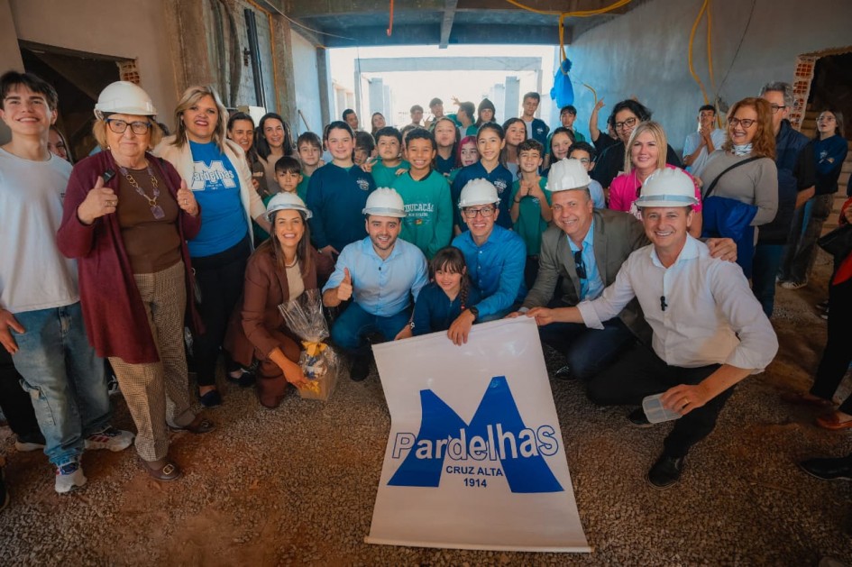 Um grupo de pessoas, incluindo adultos e crianças em uniformes escolares, está reunido posando para a foto em meio à obra na escola. Ao centro, há um banner com a logo da instituição.