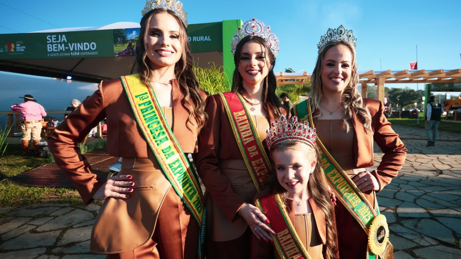 Quatro soberanas da 48ª Expointer posam sorridentes para foto ao ar livre, em frente ao pórtico de entrada do Parque de Exposições Assis Brasil, em Esteio (RS). Elas usam trajes em tom marrom e faixas coloridas que identificam seus títulos: Rainha, Primeira Princesa, Segunda Princesa e Rainha Infantil. Todas usam coroas ornamentadas. Ao fundo, há pessoas circulando pelo espaço e uma placa verde com os dizeres “Seja bem-vindo – A gente estava tchê esperando”. O céu está claro e ensolarado.