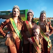 Quatro soberanas da 48ª Expointer posam sorridentes para foto ao ar livre, em frente ao pórtico de entrada do Parque de Exposições Assis Brasil, em Esteio (RS). Elas usam trajes em tom marrom e faixas coloridas que identificam seus títulos: Rainha, Primeira Princesa, Segunda Princesa e Rainha Infantil. Todas usam coroas ornamentadas. Ao fundo, há pessoas circulando pelo espaço e uma placa verde com os dizeres “Seja bem-vindo – A gente estava tchê esperando”. O céu está claro e ensolarado.