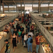 Foto geral do pavilhão. No centro do quadro, está um corredor enquadrado levemente por cima, com várias pessoas caminhando. Ao redor, os estandes, de cor branca, com produtos em exibição nas bancadas.