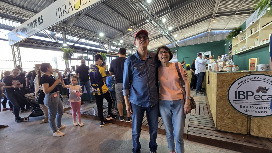 Um homem e uma mulher posam para a câmera em frente ao estante da Feira de Azeites. Diversas pessoas aparecem ao fundo, participando da feira.