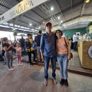 Um homem e uma mulher posam para a câmera em frente ao estante da Feira de Azeites. Diversas pessoas aparecem ao fundo, participando da feira. 