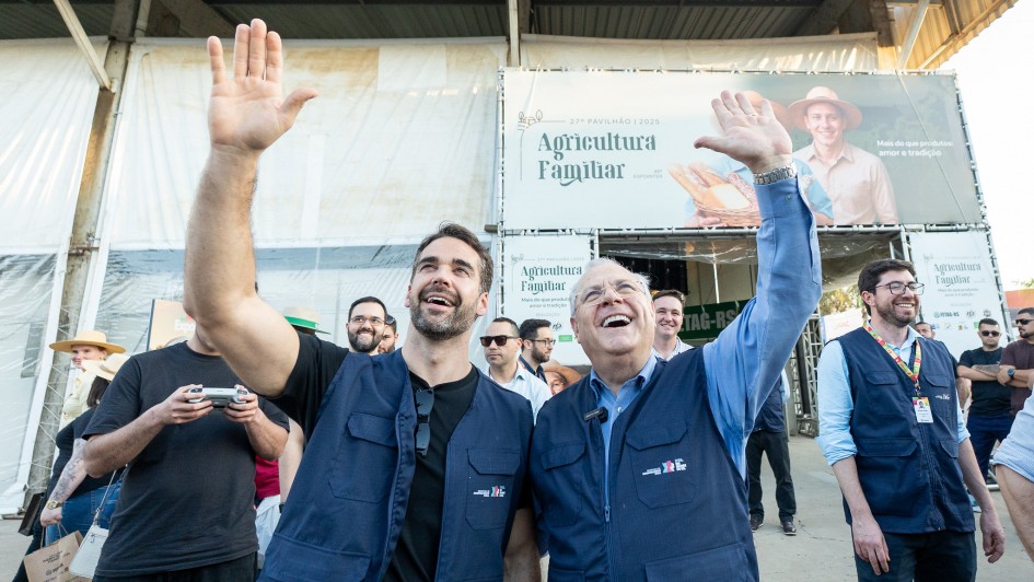 Foto em ambiente aberto. O governador está ao lado do secretário Vilson Covatti. Ambos olham para cima e acenam as mãos. Ao fundo, há diversas pessoas. Eles estão em frente ao Pavilhão da Agricultura Familiar na feira.