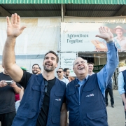 Foto em ambiente aberto. O governador está ao lado do secretário Vilson Covatti. Ambos olham para cima e acenam as mãos. Ao fundo, há diversas pessoas. Eles estão em frente ao Pavilhão da Agricultura Familiar na feira. 