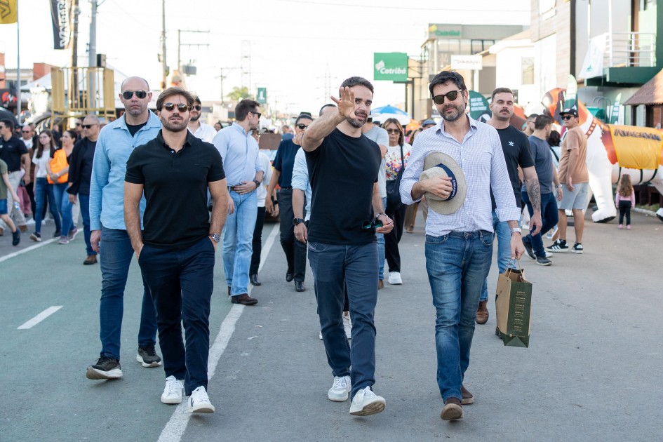 Três homens caminham pelas vias do Parque de Exposições Assis Brasil. Ao fundo, há diversas pessoas circulando pela feira. O governador está ao centro e seu marido está ao lado esquerdo da imagem.