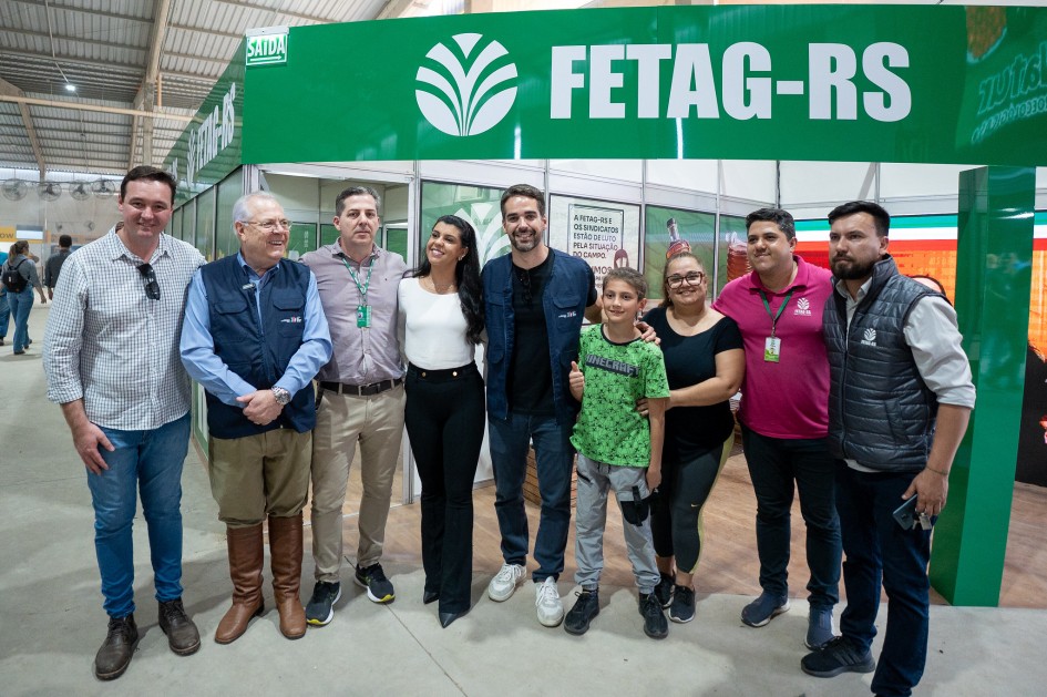 Nove pessoas estão reunidas, em pé, posando para a foto. São seis homens, duas mulheres e um menino. Eles estão em frente ao estande da Fetag, em um pavilhão da feira.