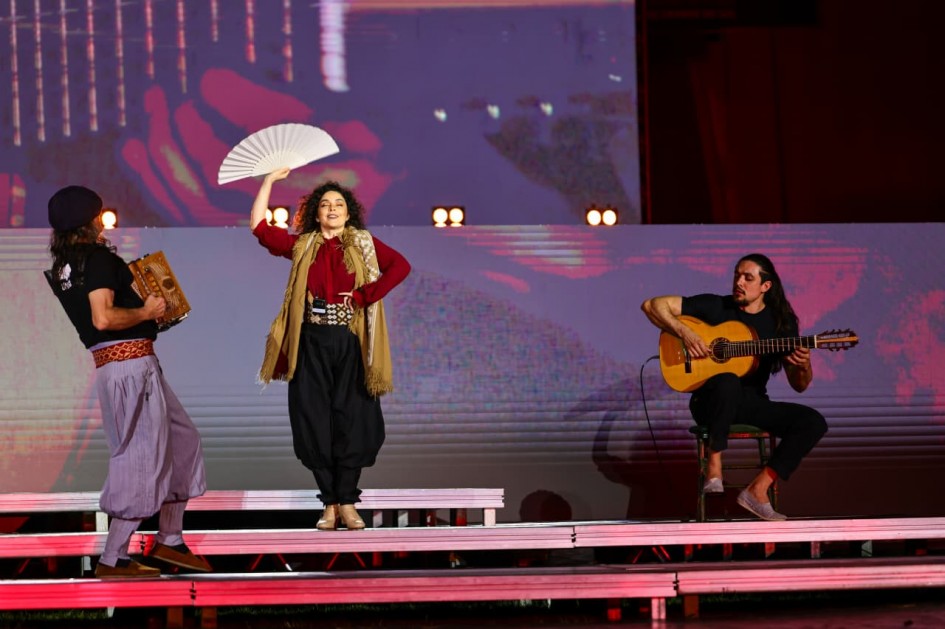 Esta é uma fotografia de três artistas em um palco, durante uma apresentação de ópera ou teatro ao ar livre.
No centro do palco, uma mulher de cabelos cacheados e escuros está em pé sobre um degrau. Ela veste uma blusa vermelha de mangas compridas, uma saia preta volumosa sobre uma calça preta e um colete de cor ocre com franjas. Ela segura um leque branco com a mão direita, com a outra mão no quadril, e sua expressão é de performance.
À sua direita, um homem de cabelo comprido e escuro está sentado em uma cadeira e toca um violão. Ele veste uma blusa preta de manga curta e calça preta.
À esquerda da mulher, um homem de gorro preto e camiseta preta, com calças listradas cinza e brancas, caminha com um instrumento de percussão parecido com um acordeão em suas mãos.
O palco é preto e a iluminação é dramática, com luzes vermelhas e roxas que iluminam o trio. Ao fundo, um grande telão exibe imagens abstratas em tons de vermelho e branco.