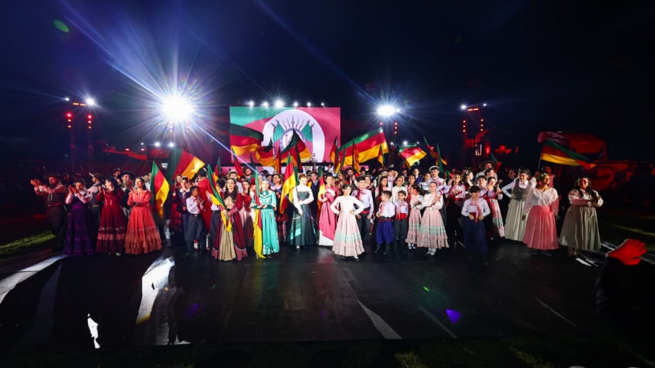 Fotografia de um espetáculo cultural realizado à noite em palco aberto. No centro, dezenas de artistas, incluindo homens, mulheres e crianças, vestem trajes típicos gaúchos coloridos. Muitos carregam bandeiras do Rio Grande do Sul erguidas, compondo uma cena vibrante e festiva. Ao fundo, um telão exibe a bandeira do Estado, iluminada por refletores intensos que criam feixes de luz sobre o grupo. O conjunto transmite clima de celebração, tradição e identidade cultural gaúcha.