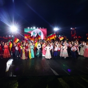 Fotografia de um espetáculo cultural realizado à noite em palco aberto. No centro, dezenas de artistas, incluindo homens, mulheres e crianças, vestem trajes típicos gaúchos coloridos. Muitos carregam bandeiras do Rio Grande do Sul erguidas, compondo uma cena vibrante e festiva. Ao fundo, um telão exibe a bandeira do Estado, iluminada por refletores intensos que criam feixes de luz sobre o grupo. O conjunto transmite clima de celebração, tradição e identidade cultural gaúcha.