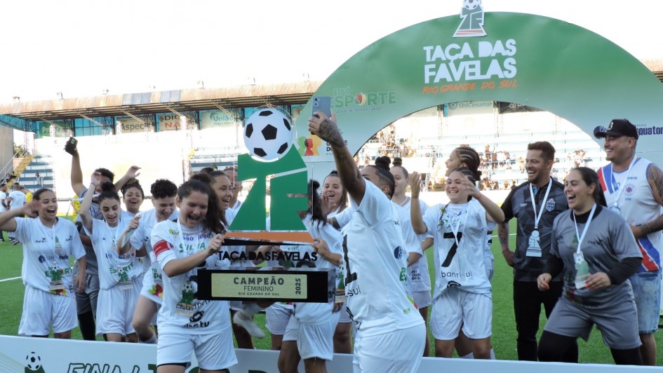 A imagem mostra um grupo de jogadoras de futebol comemorando a conquista da Taça das Favelas 2025 em um campo ao ar livre. Elas estão vestidas com uniformes brancos e seguram um grande troféu.
