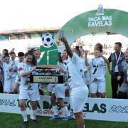 A imagem mostra um grupo de jogadoras de futebol comemorando a conquista da Taça das Favelas 2025 em um campo ao ar livre. Elas estão vestidas com uniformes brancos e seguram um grande troféu.