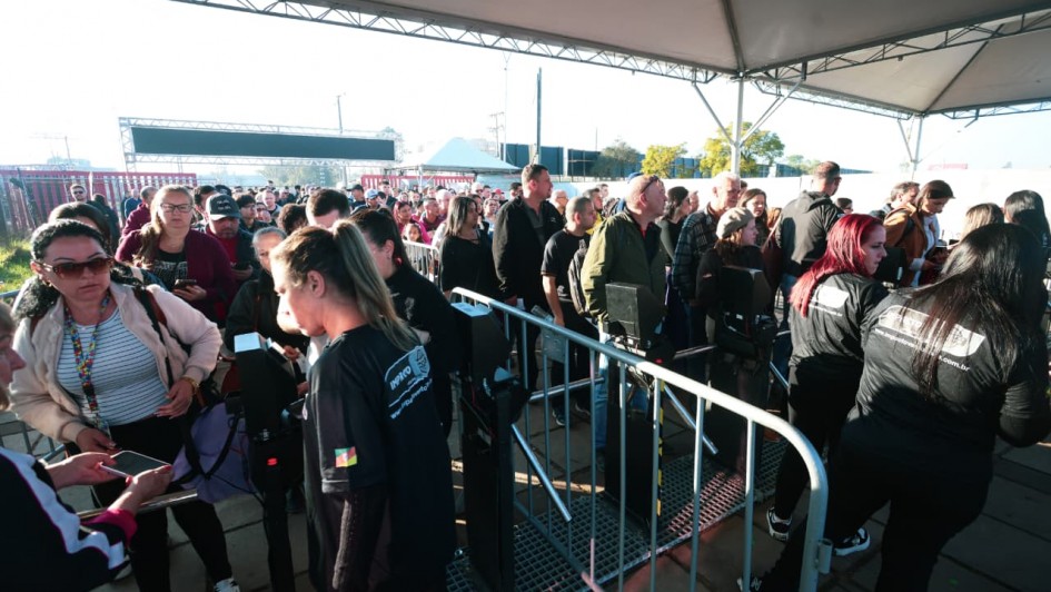 Fila de visitantes na entrada da 48ª Expointer, em Esteio (RS). A imagem mostra um grande grupo de pessoas passando pela catraca de acesso, sob uma cobertura metálica. Funcionários, vestidos com camisetas pretas, realizam a conferência dos ingressos nos celulares do público. Ao fundo, mais pessoas aguardam na fila para entrar no parque de exposições. O ambiente está iluminado pela luz do sol da manhã.