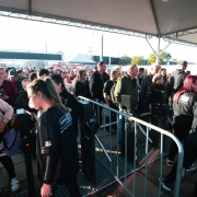 Fila de visitantes na entrada da 48ª Expointer, em Esteio (RS). A imagem mostra um grande grupo de pessoas passando pela catraca de acesso, sob uma cobertura metálica. Funcionários, vestidos com camisetas pretas, realizam a conferência dos ingressos nos celulares do público. Ao fundo, mais pessoas aguardam na fila para entrar no parque de exposições. O ambiente está iluminado pela luz do sol da manhã.