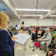 Mulher loira, de óculos, fala ao microfone em um auditório lotado. Ela está de costas para a câmera, lendo de uma folha sobre um púlpito transparente. À frente, diversas pessoas estão sentadas, entre elas adultos, idosos e uma criança sentada no chão. No palco, um homem está sentado à mesa, observando a apresentação. O ambiente é iluminado e possui teto branco com projetor instalado.