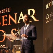 Foto em plano americano de Leite discursando no púlpito. Ao fundo, um grande painel preto com os dizeres em dourado: Troféu Senar o Sul.