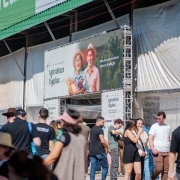 A imagem mostra um evento ao ar livre com várias pessoas caminhando e interagindo em frente a um grande estande. Na parte superior do estande há uma placa verde com o logotipo do Sicredi. Abaixo, há um banner grande com a foto de duas pessoas sorridentes (um homem e uma mulher mais velhos, com roupas típicas de agricultores), segurando cestas com produtos agrícolas como pão, uvas e verduras. No banner está escrito:

"29ª Expointer 2025
Agricultura Familiar
Mais do que produtos: amor e tradição".

Muitas pessoas estão passando ou paradas em frente ao estande. A maioria veste roupas casuais de verão e alguns usam chapéus para se proteger do sol. O ambiente parece ser uma feira ou exposição agropecuária, com clima descontraído e ensolarado.