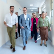 A imagem mostra um grupo de pessoas caminhando por um corredor bem iluminado, aparentemente em uma instituição de saúde. Na frente do grupo, há quatro pessoas visivelmente em destaque:

À esquerda, um homem de barba, vestindo camisa branca de mangas dobradas, calça verde e sapatos marrons. Ele segura alguns papéis na mão direita.

Ao seu lado, um homem com barba grisalha e óculos, vestindo um terno azul-claro com padrão xadrez e sapatos marrons. Ele está com as mãos nos bolsos.

À direita, uma mulher mais velha, com cabelos grisalhos presos, vestindo uma blusa verde, saia marrom e sapatos verdes. Ela tem um crachá pendurado no pescoço e carrega uma pequena bolsa verde a tiracolo.

Atrás dela, há outra mulher de cabelos loiros, usando óculos e um casaco rosa-choque sobre uma roupa escura. Ela também usa crachá.

Ao fundo, outras pessoas acompanham a caminhada. O ambiente é limpo e claro, com paredes brancas, janelas grandes à direita e piso cinza-claro.