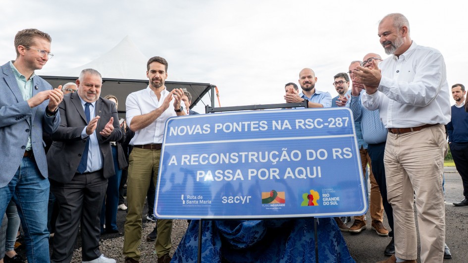 Imagem mostra o governador Eduardo Leite e outras autoridades aplaudindo ato. Eles estão em frente a uma placa azul com os dizeres: "Novas pontes na RSC-287: A reconstrução do RS passa por aqui", com logos do governo do estado e da empresa concessionária da rodovia.