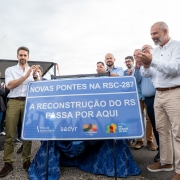 Imagem mostra o governador Eduardo Leite e outras autoridades aplaudindo ato. Eles estão em frente a uma placa azul com os dizeres: "Novas pontes na RSC-287: A reconstrução do RS passa por aqui", com logos do governo do estado e da empresa concessionária da rodovia.