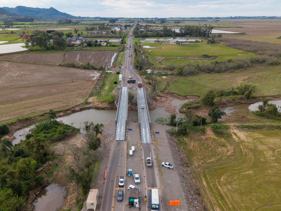 Foto aérea mostra a rodovia em extensão, com duas pontes metálicas em primeiro plano, sobre o arroio. Nas laterais da rodovia, campos e lavouras. Ao fundo, o céu e uma montanha ao fundo.