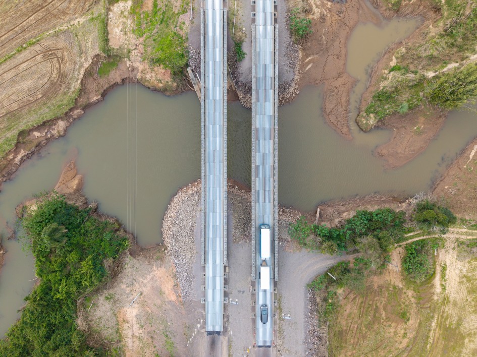 Foto aérea mostra em detalhe a ponte sobre o Arroio Grande, destacando a ponte metálica e mostrando o leito do rio, barrento, em forma irregular, com vegetação à volta. Sobre a ponte começam a passar dois veículos grandes, possivelmente caminhões.