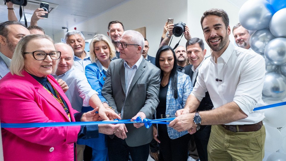 A imagem mostra um grupo de pessoas participando de uma cerimônia de inauguração. No centro, várias mãos seguram uma tesoura para cortar uma fita azul, simbolizando a abertura oficial do espaço. Ao fundo, outras pessoas acompanham o momento, algumas registrando a cena com câmeras e celulares. Há balões prateados e brancos decorando o ambiente, reforçando o clima festivo do evento.