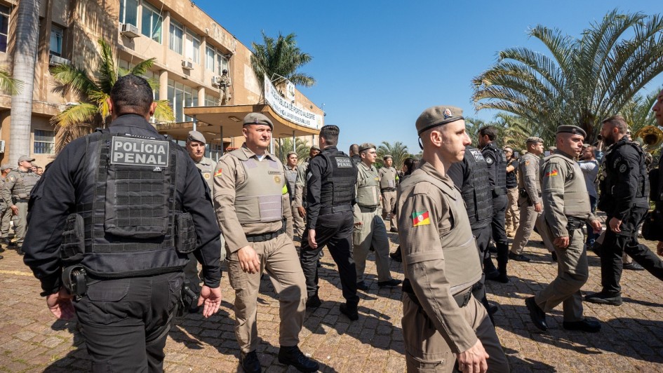 Foto mostra fileiras de policiais que se cruzam em alinhamento. À esquerda da foto, está o prédio da Cadeia Pública de Porto Alegre, com fachada em amarelo e bege. Marcham em direção ao prédio, de costa para a foto, os policiais penais, com uniformes pretos. Em alguns é possível ver nas costas o emblema da Polícia Penal. No outro sentido, em direção ao jardim, marcham os policiais militares, com uniformes beges e boinas também beges.