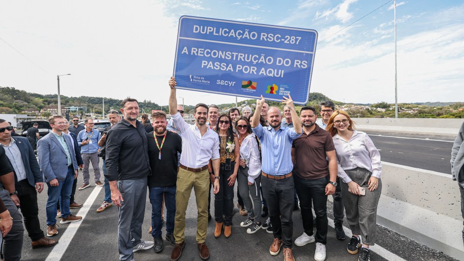 A imagem mostra um grupo de pessoas reunidas sobre uma rodovia. No centro, duas pessoas seguram uma placa azul com os dizeres: "Duplicação RSC-287 - A reconstrução do RS passa por aqui".