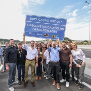 A imagem mostra um grupo de pessoas reunidas sobre uma rodovia. No centro, duas pessoas seguram uma placa azul com os dizeres: "Duplicação RSC-287 - A reconstrução do RS passa por aqui".
