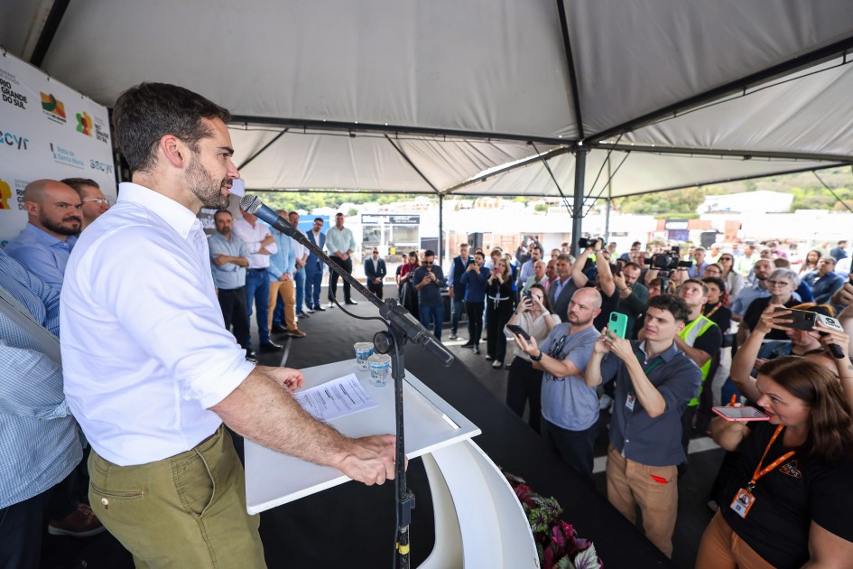 Governador Eduardo Leite fala ao microfone em um evento ao ar livre, sob uma tenda. Há uma plateia em pé, algumas pessoas tirando fotos. O fundo tem logotipos de empresas e órgãos públicos, sugerindo que é um evento oficial.