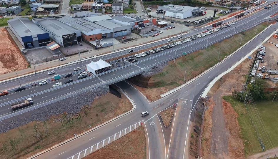 Rodovia larga com vários acessos e saídas, cercada por áreas verdes, morros e construções. Há movimento de carros e sinais de urbanização misturada com natureza.