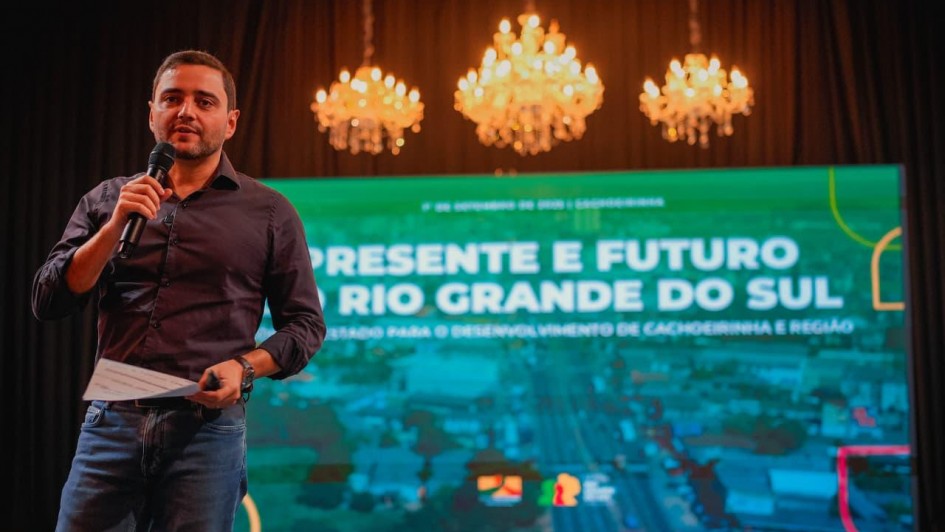 Um homem está em pé sobre um palco, segurando um microfone com uma mão e uma folha de papel com a outra. Ele veste uma camisa preta e jeans. Ao fundo, há um grande telão com os dizeres: "Presente e Futuro do Rio Grande do Sul – Estratégias para o desenvolvimento de Cachoeirinha e região". No topo do telão, está a data do evento: "1º de setembro de 2023 | Cachoeirinha". Acima da tela, três lustres de cristal iluminam o ambiente, sugerindo que o evento ocorre em um teatro ou auditório. O clima da imagem é formal, com foco em uma apresentação institucional.