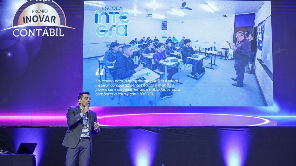 Um homem de terno cinza e gravata azul segura um microfone e fala no palco durante evento. Atrás dele, um telão exibe o logotipo “Prêmio Inovar Contábil” e a imagem de uma sala de aula do projeto Escola Íntegra, com estudantes sentados em carteiras enquanto um professor ministra a aula. No telão, aparece também a frase: “Educação para a integridade pública é sobre inspirar comportamentos éticos e munir os jovens com conhecimentos e habilidades para combater a corrupção” (OCDE).