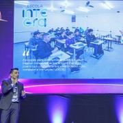 Um homem de terno cinza e gravata azul segura um microfone e fala no palco durante evento. Atrás dele, um telão exibe o logotipo “Prêmio Inovar Contábil” e a imagem de uma sala de aula do projeto Escola Íntegra, com estudantes sentados em carteiras enquanto um professor ministra a aula. No telão, aparece também a frase: “Educação para a integridade pública é sobre inspirar comportamentos éticos e munir os jovens com conhecimentos e habilidades para combater a corrupção” (OCDE).