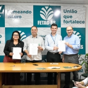 A imagem mostra quatro pessoas em pé atrás de uma mesa de madeira, segurando documentos assinados e sorrindo para a câmera. Eles estão participando de um evento formal relacionado à agricultura familiar. Ao fundo, há um painel com logotipos e frases institucionais, como "FETAG-RS", "União que fortalece", "Agricultura e pecuária familiar é vida, é futuro, é alimentação" e "O futuro começa aqui".

As quatro pessoas no centro da imagem são três homens e uma mulher. A mulher está à esquerda, vestindo um blazer preto, óculos e saia vermelha com estampa. À direita dela, os três homens estão vestidos com camisas sociais, um deles com crachá. Todos seguram folhas de papel, aparentemente contratos ou acordos assinados.

À direita da imagem, uma mulher loira está sentada e concentrada em um celular. Há uma planta decorativa ao fundo e parte de uma parede de tijolos aparentes à direita.