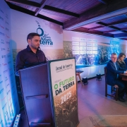Foto de de ambiente fechado, de coloração azul, com Gabriel, à esquerda, discursando junto ao púlpito. À direita, há uma mesa com várias pessoas sentadas em fila.