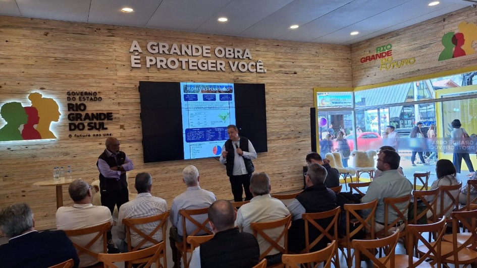 Foto de uma sala dentro do Parque Assis Brasil, tirada do fundo. Um homem aparece de pé, falando ao microfone, com várias pessoas assistindo sentadas. Há uma tela com apresentação atrás dele, e uma logo do governo na parede.
