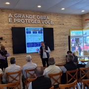 Foto de uma sala dentro do Parque Assis Brasil, tirada do fundo. Um homem aparece de pé, falando ao microfone, com várias pessoas assistindo sentadas. Há uma tela com apresentação atrás dele, e uma logo do governo na parede.