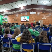 Em uma sala, diversos estudantes estão reunidos, sentados em cadeiras, para assistir a live projetada em uma TV no centro da imagem. Os estudantes estão de costas e na tela aparece o governador Eduardo Leite falando. 