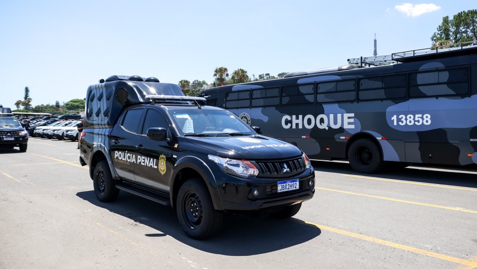 Foto mostra uma viatura da Polícia Penal em primeiro plano, na cor preta, com emblema em amarelo. Ao fundo, há um ônibus, com estampa camuflada azul e cinza, com a palavra "choque" em destaque na cor branca.